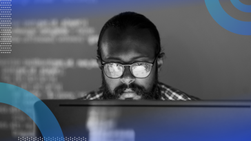 Cryptanalysis image of a man sitting in front of a computer screen. We are looking at the man head-on from behind the computer monitor. The computer screen reflects in the man's thick, dark glasses.