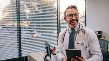 A male doctor smiles while looking off camera and holds a tablet