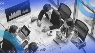 A group of people sit around a conference table with graphs and monitors showing statistics