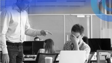 A person is sitting at a desk covering their eyes while a manager, who is standing, is pointing at them.