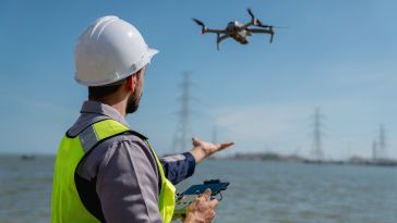 A person wearing a hard hat and bright green safety vest stands outside next to the water and holds a controller in their hand while a drone flies in the air nearby.