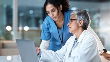 A medical professional in a white lab coat sits at a desk in front of a laptop computer while another professional wearing blue scrubs stands over their shoulder looking at the screen.