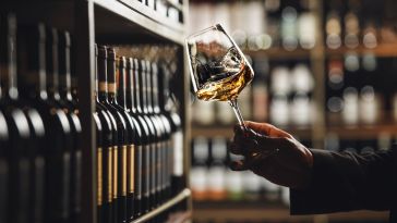 A close up of a hand holding a glass of white wine in front of a shelf filled with wine bottles.