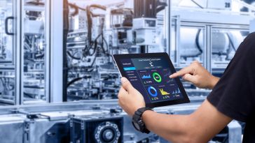 A pair of hands holds a tablet in front of a room full of industrial machinery.