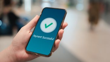 A close up of a hand holding a mobile phone with a green check mark on the screen and text stating "Payment Successful."