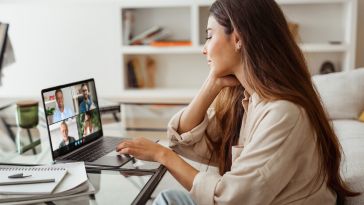 A woman in a home office sits in front of a laptop with a video conference call featuring four other professionals on the screen.